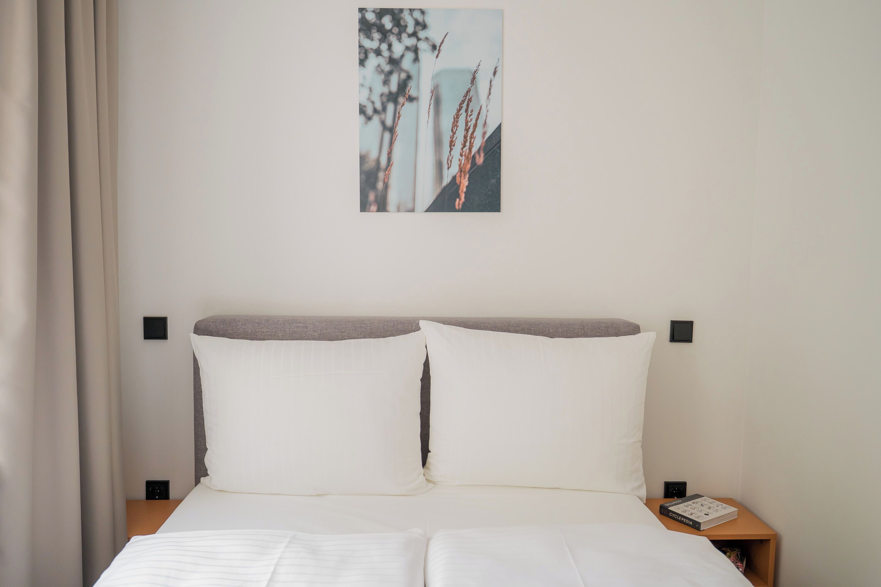 A neatly made bed with white linens, a headboard with a grey panel, and a small bedside table with a book and a power outlet.