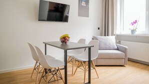 A modern living room with a dining table, white chairs, a grey sofa, a flat-screen TV, and a window with a view. - Homaris Apartments Nibelungen (Frankfurt)