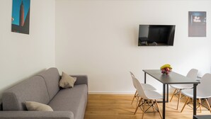 A modern living room with a grey sofa, a dining table with white chairs, a flat-screen TV mounted on the wall, and a framed picture of a building. - Homaris Apartments Nibelungen (Frankfurt)