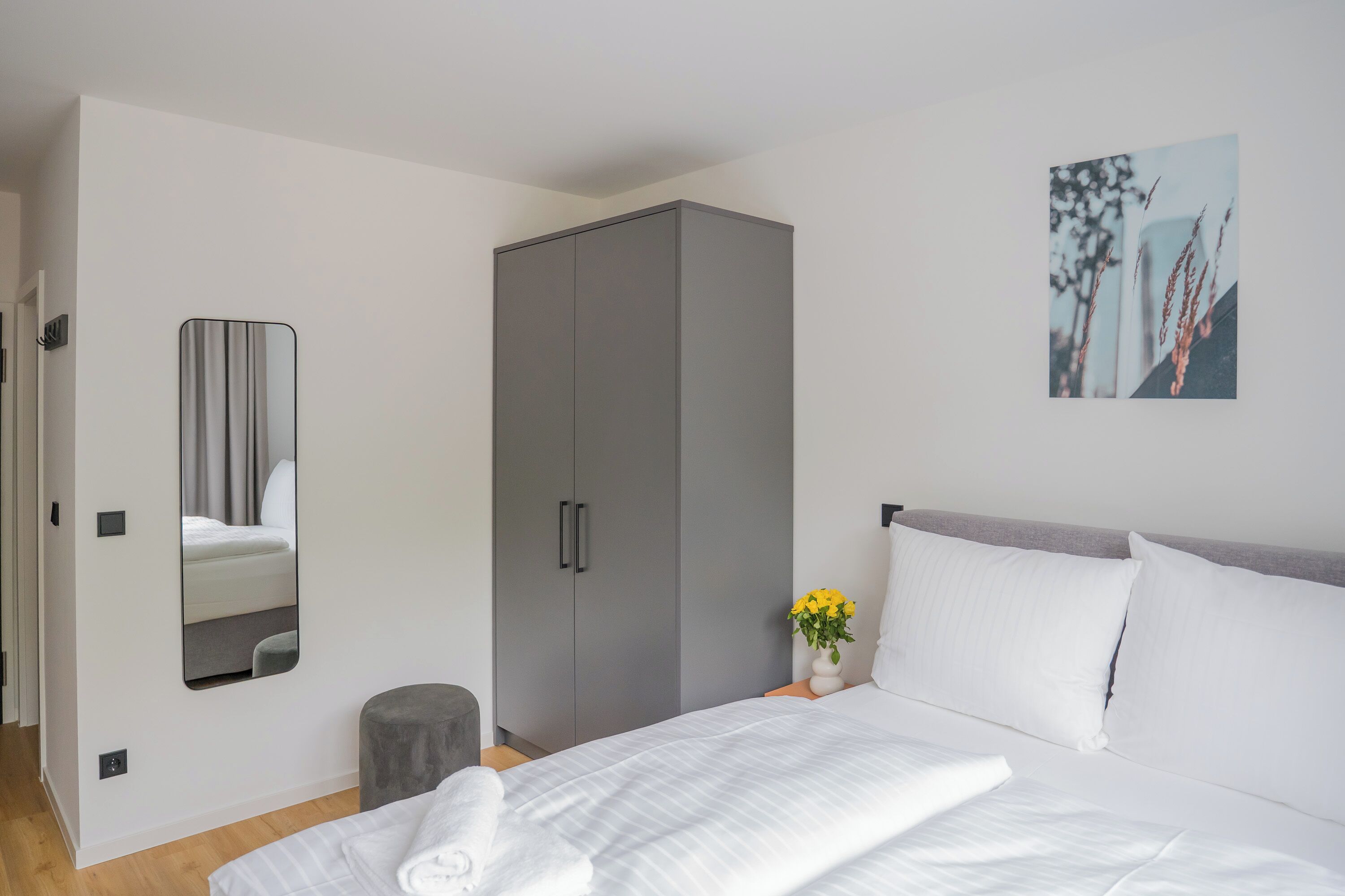 Un dormitorio moderno con una cama grande, un armario gris, un taburete pequeño y un espejo montado en la pared.