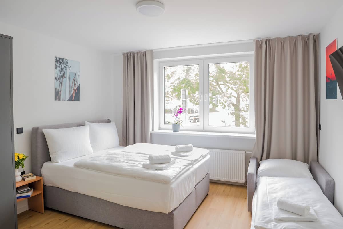 Una habitación de hotel moderna con una cama grande, una cama más pequeña, una ventana con cortinas, un radiador y una pequeña mesa con un jarrón de flores.