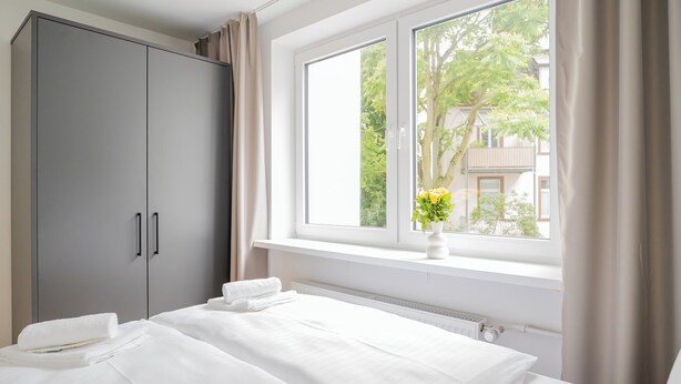 A hotel room with a large wardrobe, a bed with white linens, a window with a view of buildings and trees, and a vase with flowers on the windowsill. - Homaris Apartments Nibelungen (Frankfurt)