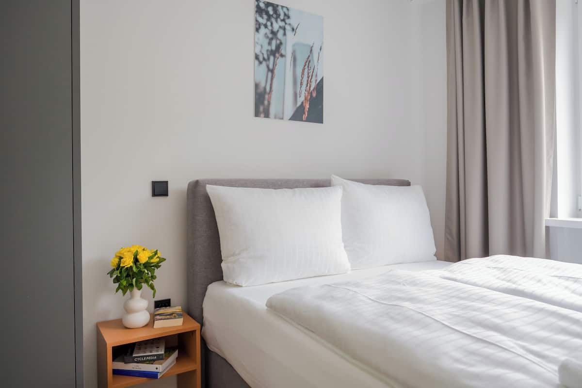 Una cama bien hecha con sábanas blancas, una mesita de noche con un jarrón de flores amarillas y una obra enmarcada en la pared.