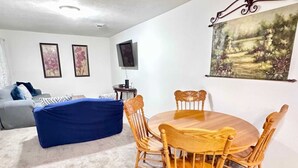 A living room with a blue sofa, a wooden dining table with chairs, a TV mounted on the wall, and a painting above the table. - Entire Spacious 4 BR Home for Teams (Port Arthur)