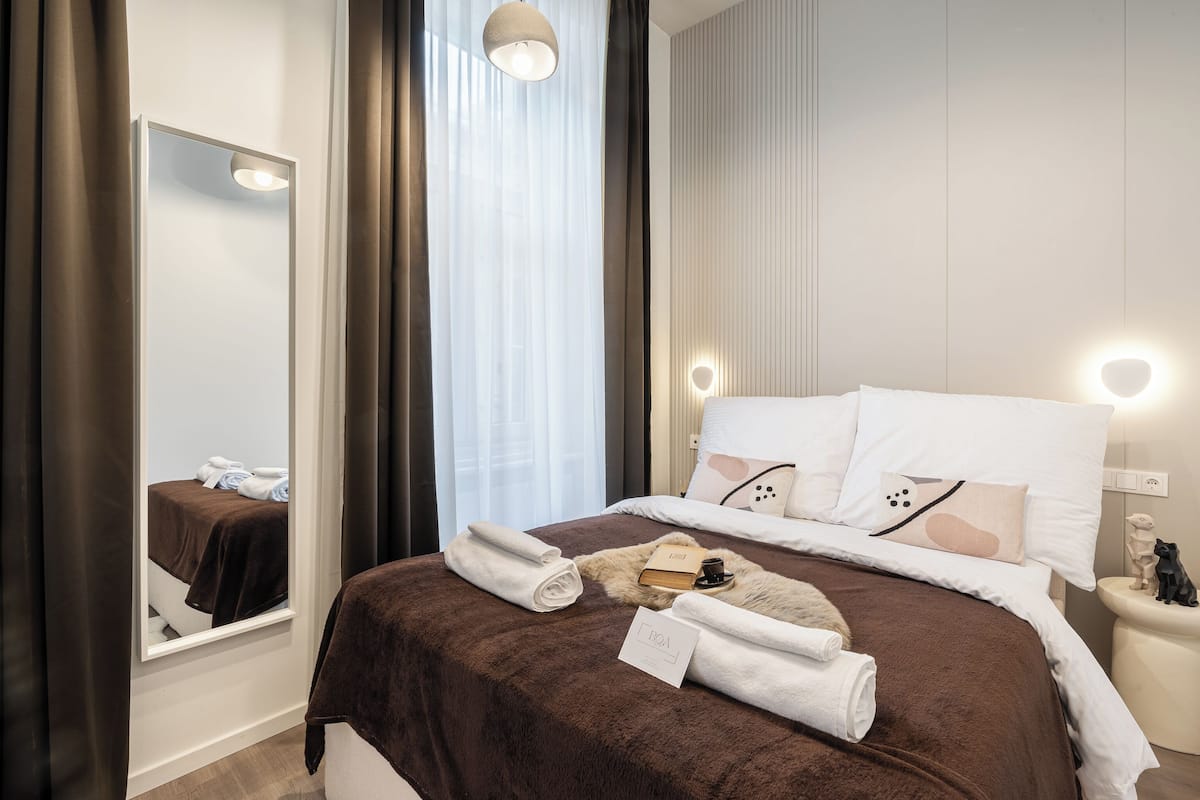 Un lit bien fait, avec une couverture marron, des oreillers blancs et un petit livre. On y voit une table de chevet avec une lampe et un objet décoratif. Un miroir reflète une partie de la pièce.