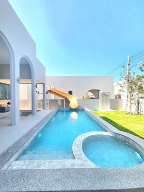 A swimming pool with a slide, surrounded by a modern building with arched walkways and a clear sky. - Phattra Pool villa Cha-am (Cha-am)