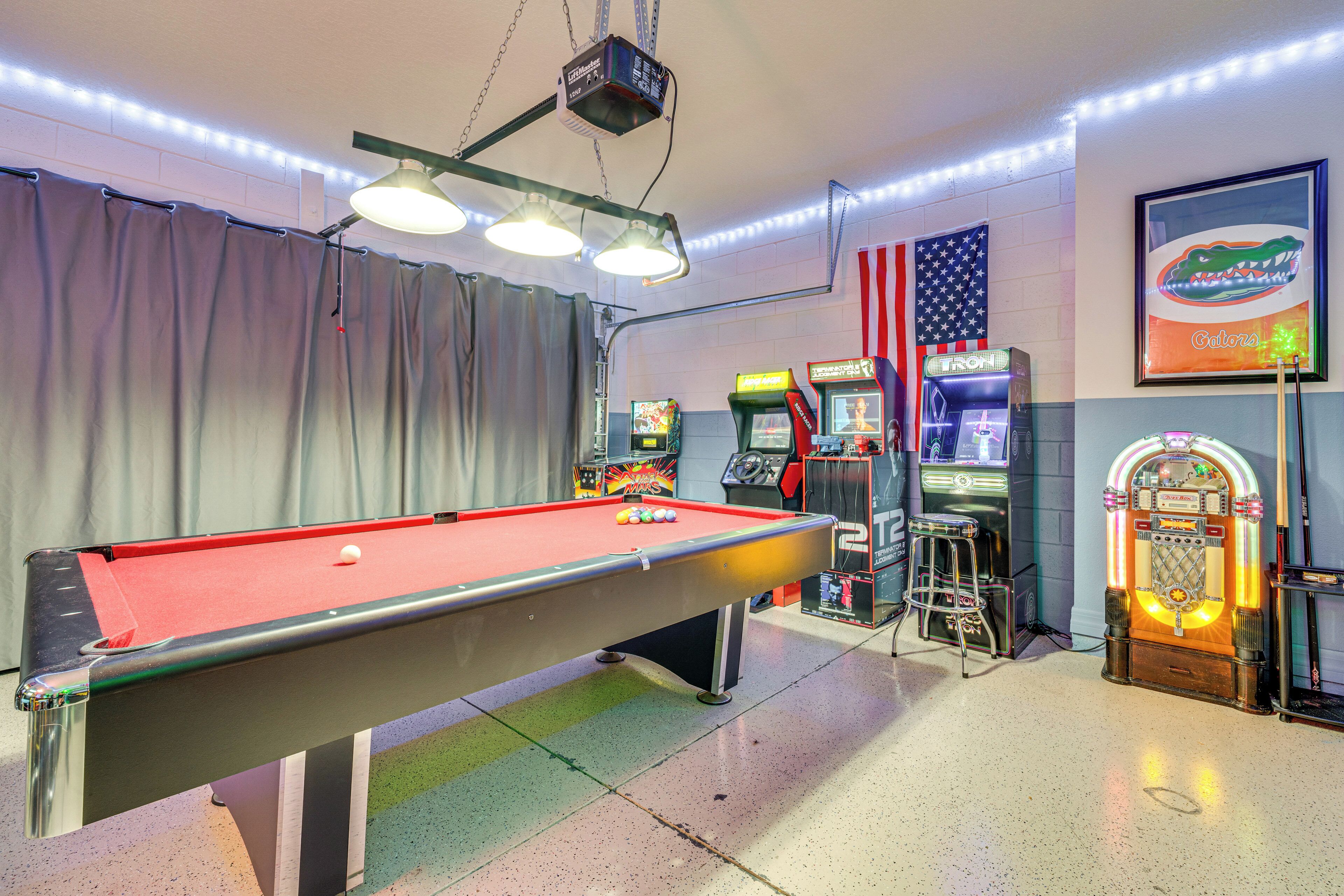 A pool table with lights, arcade machines, and a jukebox in a room with a striped floor and curtains.
