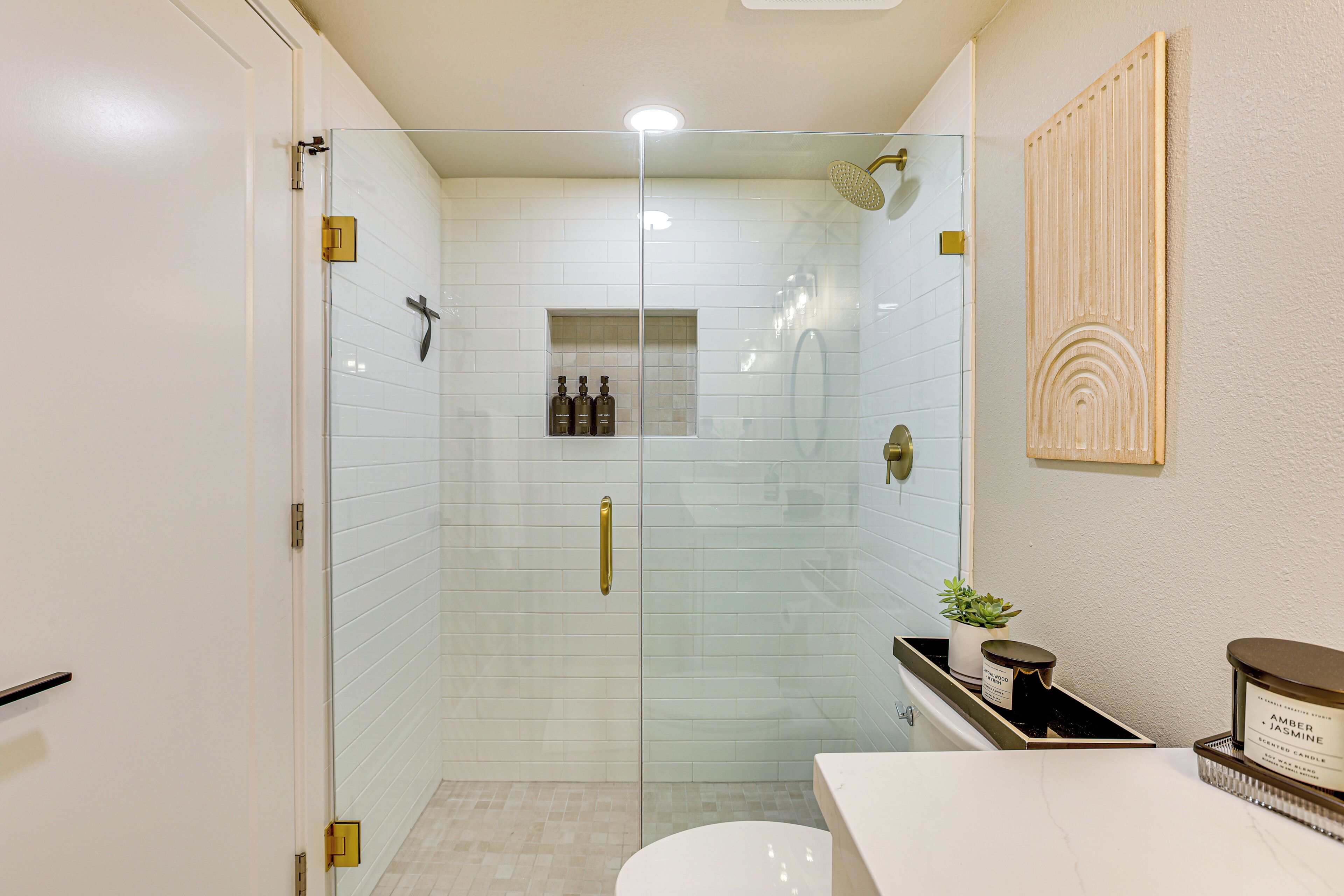 A modern bathroom with a glass-enclosed shower, white tiled walls, a wooden panel wall art piece, and a small shelf with toiletries.