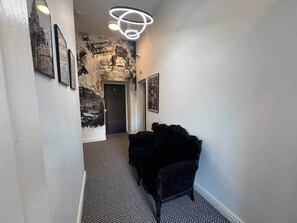 Un couloir avec un tapis à motifs noir et blanc, une chaise noire et un mur orné de tableaux encadrés et d’une fresque.