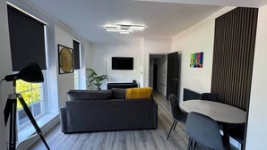 Un saló modern amb un sofà gris, una taula de menjador amb caires, un televisor penjat a la paret i una planta.