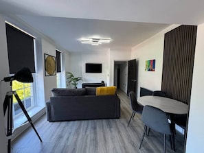 Ein modernes Wohnzimmer mit einem grauen Sofa, einem Esstisch mit Stühlen, einem an der Wand befestigten Fernseher und einer Pflanze.