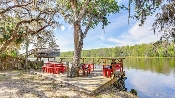 A wooden dock with red chairs and tables overlooking a calm lake, surrounded by trees and a wooden deck. - Dock & Fire Pit: Scenic Studio on Suwannee River! (Chiefland)