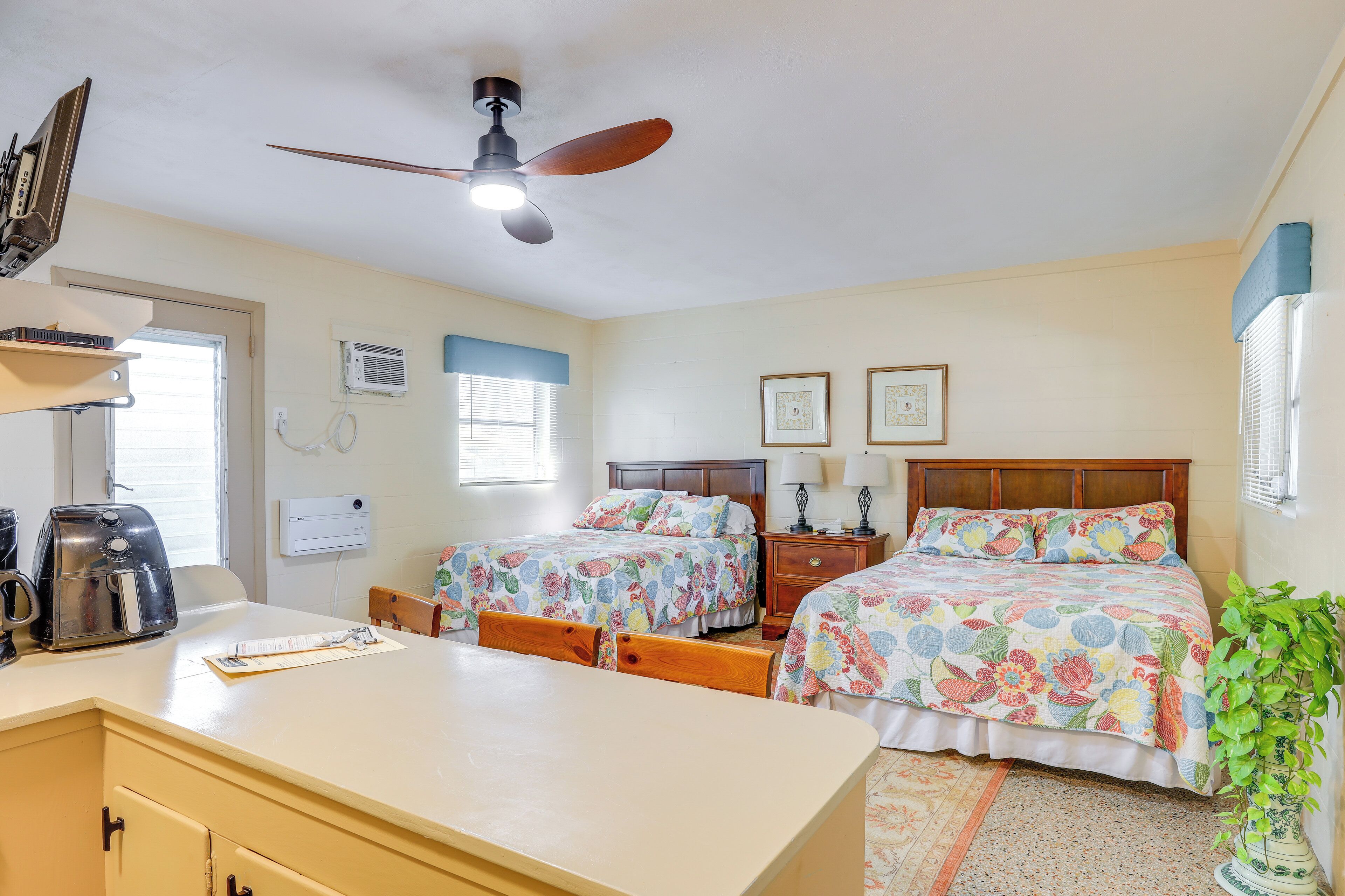 A bedroom with two beds, a ceiling fan, a bedside table with lamps, a nightstand, a dresser, a coffee maker, and a potted plant.