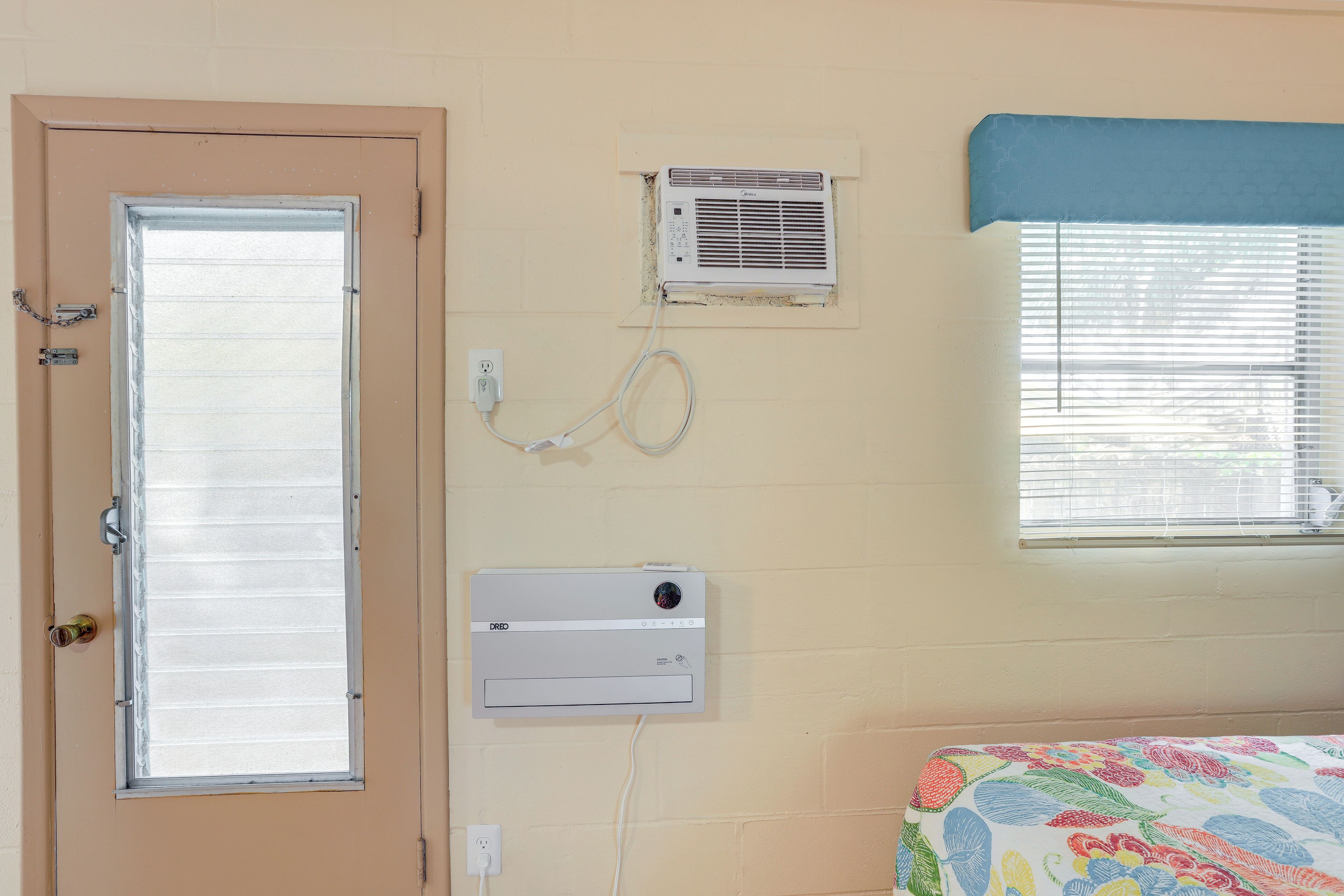 A room with a door, a window with blinds, an air conditioning unit, and a bed with a floral bedspread.