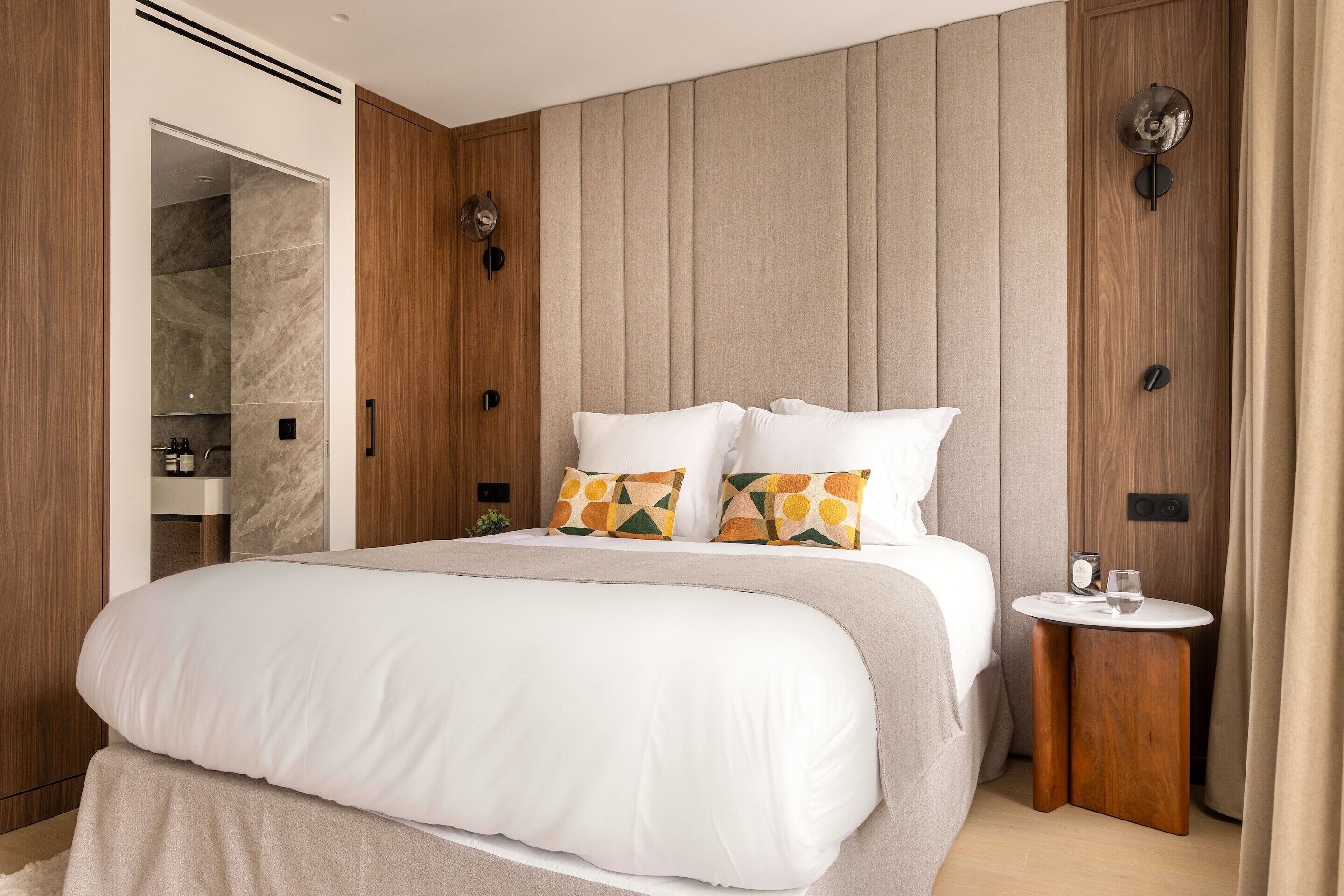 A modern bedroom with a large bed, a bedside table with a glass, and a wooden headboard.