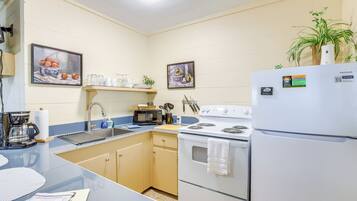 A kitchen with a sink, stove, microwave, and refrigerator. There are two framed pictures on the wall, a plant on the refrigerator, and a countertop with a coffee maker and a menu.