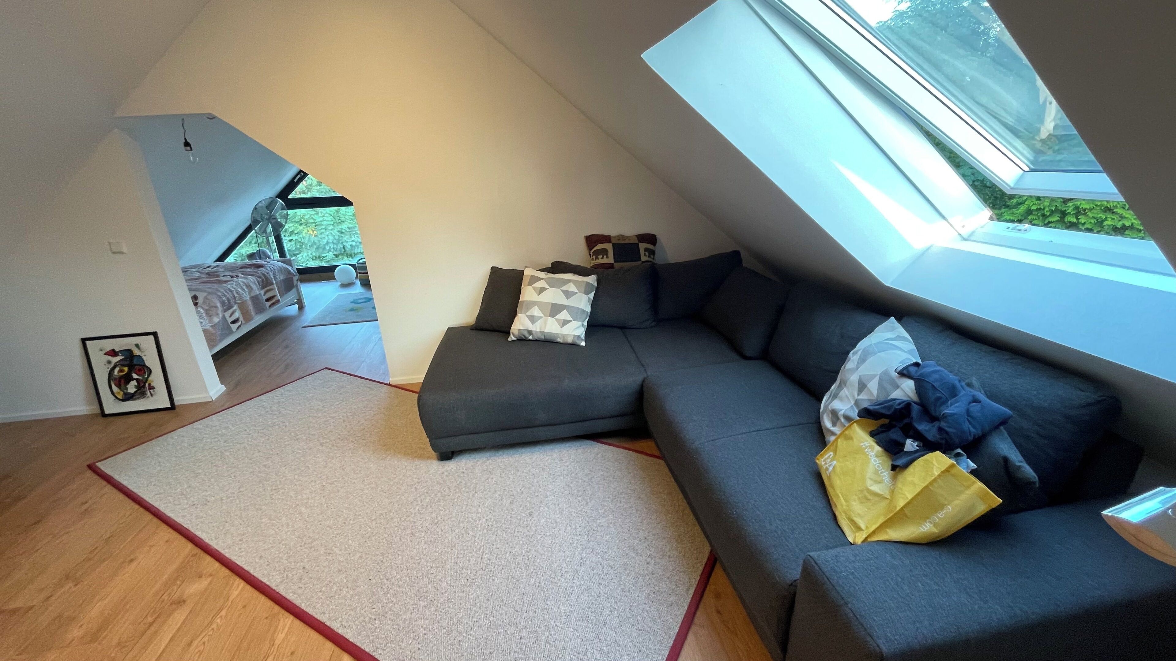 A modern loft bedroom with a sloped ceiling, a large window, a gray sofa, and a bed with a view of greenery outside.