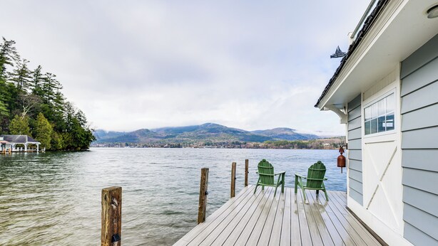 Un pontile in legno con due poltrone Adirondack verdi rivolte verso un lago, con montagne sullo sfondo.