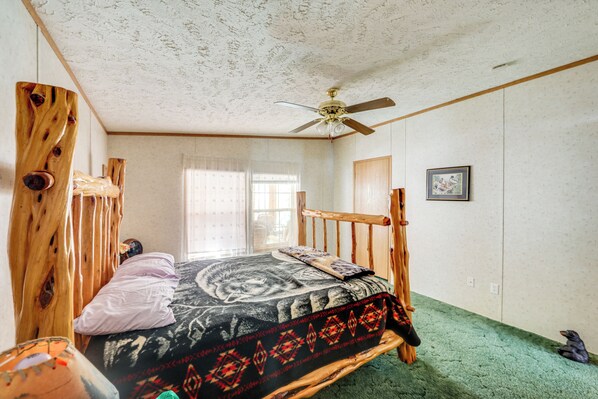 A bedroom with a wooden bed frame, a ceiling fan, a window with blinds, and a green carpet. - Creekside Retreat in the Adirondacks: Swim & Hike! (Forestport)