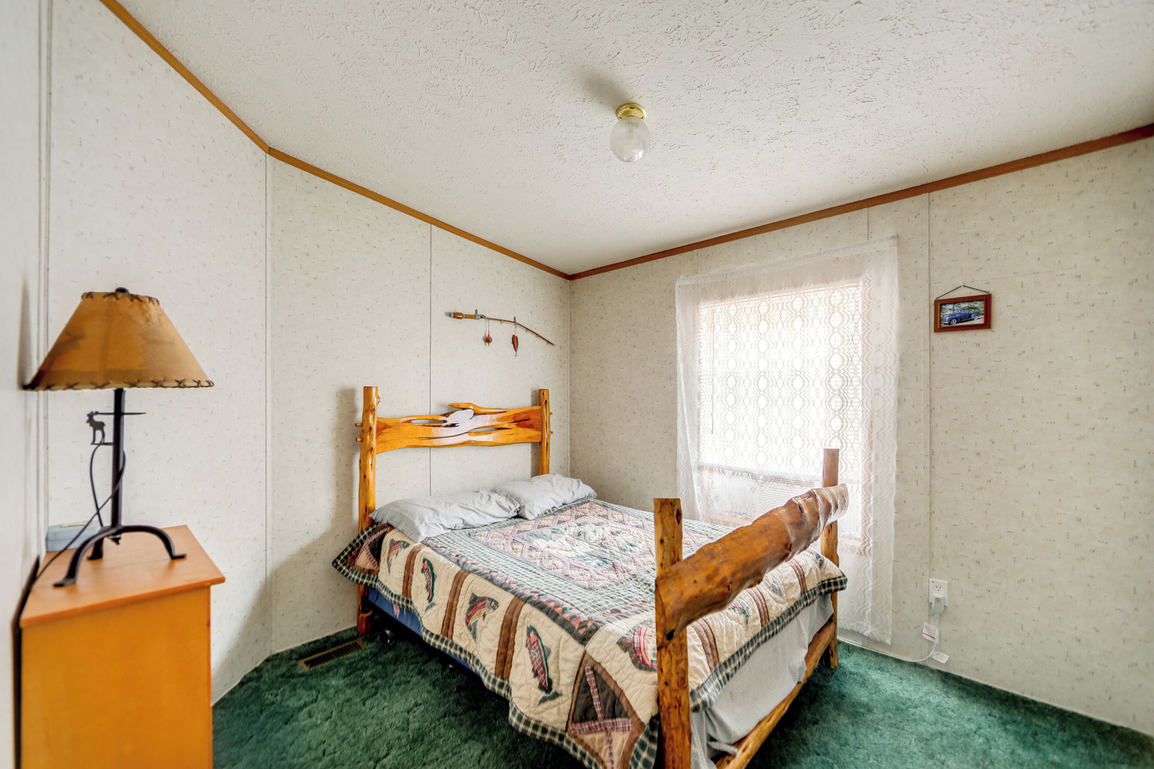 A bedroom with a wooden bed, a green carpet, a lamp, and a window with lace curtains.