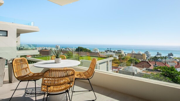 A balcony with a round table and wicker chairs, overlooking a coastal cityscape.