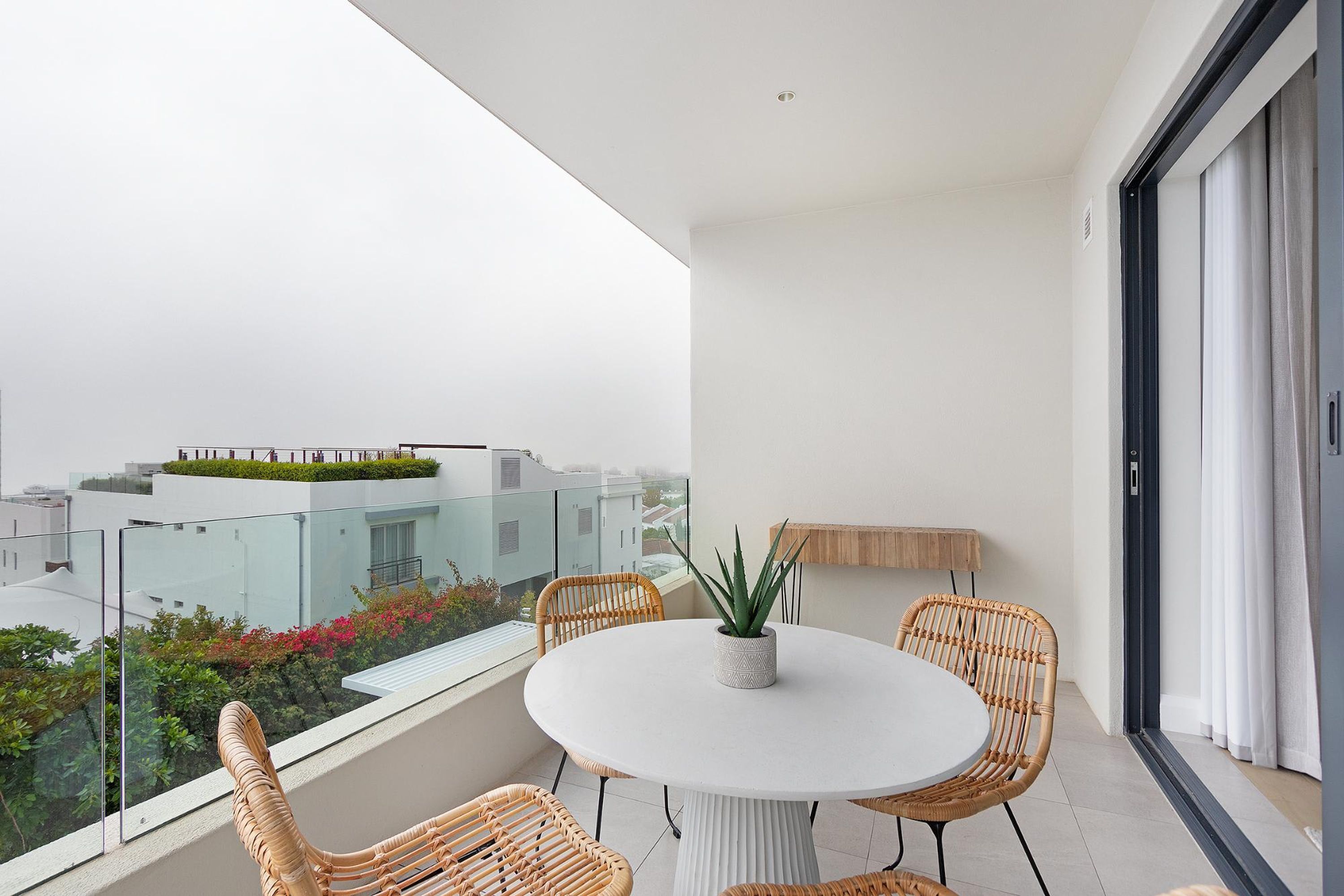 A balcony with a round table and wicker chairs, a potted plant, and a view of buildings and greenery.