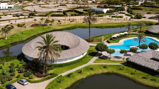 Ein Resort mit einem kreisförmigen Pool, Palmen und einem Strand im Hintergrund.