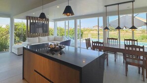 Eine moderne Küche mit einer großen Kochinsel, Pendelleuchten und Blick auf Pool und Garten.