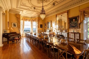Uma sala de jantar imponente com uma mesa comprida, cadeiras, um piano e um lustre.
