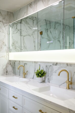 A bathroom with marble walls, a large mirror, two sinks with gold faucets, and a small potted plant. - Cozy Family Retreat With Fireplace Spacious Yard (Duarte)