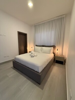 A modern bedroom with a large bed, a wooden door, a bedside table with a lamp, and a window with sheer curtains. - 3BR Villa by Vistacana Rentals (Punta Cana)
