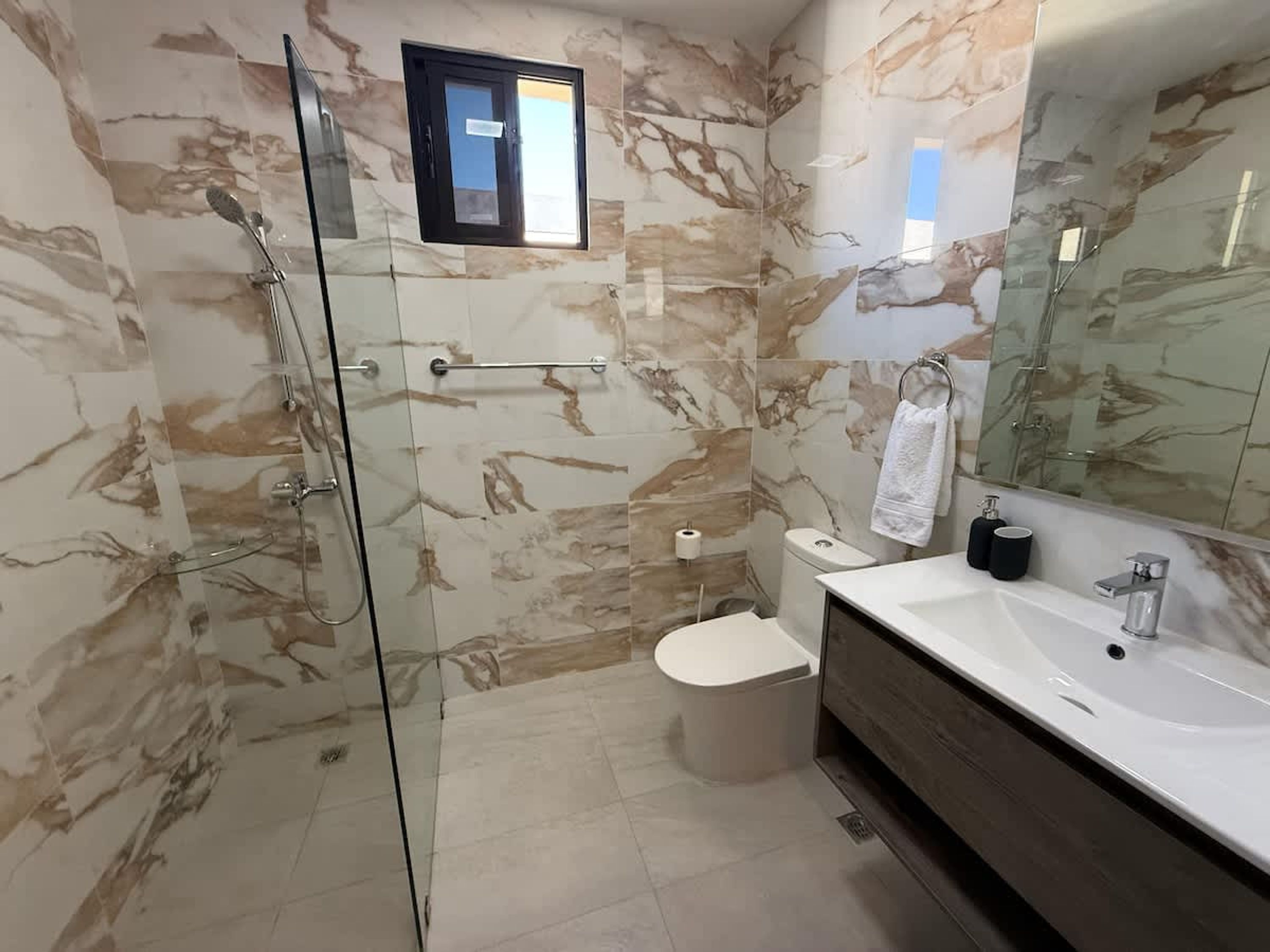 Une salle de bain moderne dotée d’une douche à parois de verre, de murs en marbre, de toilettes et d’un meuble-lavabo avec un grand miroir.