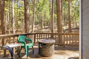 A deck with a stone fire pit, a table, and two chairs, surrounded by a forest. - Hike, Ski & Sip: Arnold Cabin 4 Mi to State Park! (Arnold)