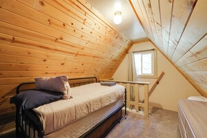 Una habitación pequeña y acogedora con paredes de madera y techo inclinado. Tiene una cama individual con colchón y almohadas, una mesita y un ventanal que proporciona luz natural.