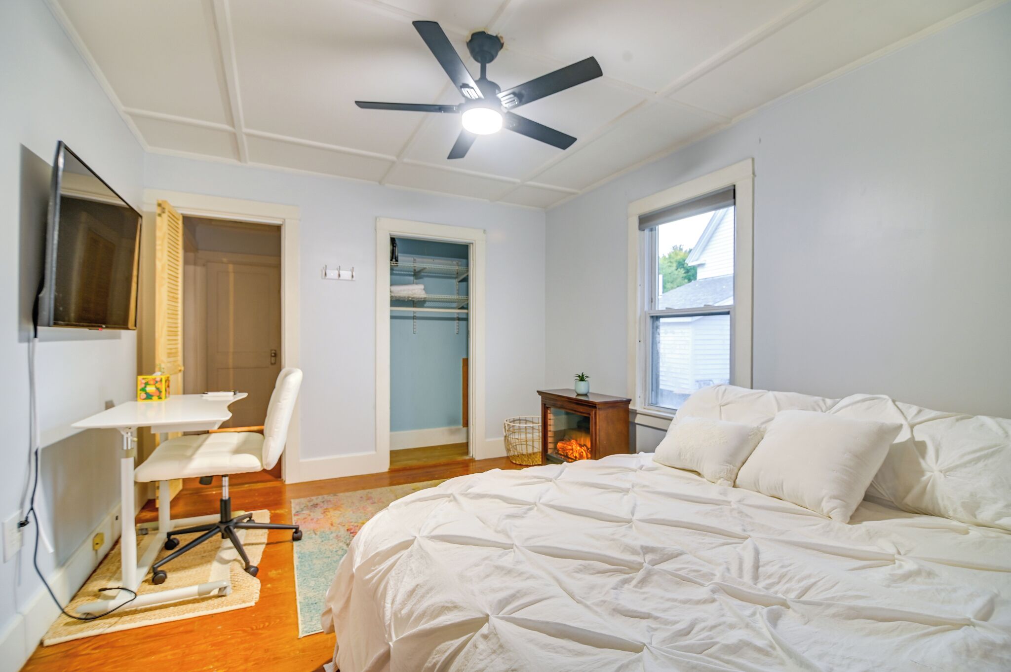 A bedroom with a bed, a desk, a chair, a ceiling fan, and a window.