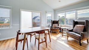A spacious room with a wooden dining table, two chairs, a leather sofa, and a small round table. Large windows provide views of the outside. - Steps to Beach: Centrally Located OBX Home! (Kill Devil Hills)