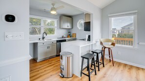 A modern kitchen with white cabinetry, a central island, bar stools, a ceiling fan, and a view of the outdoors through large windows. - Steps to Beach: Centrally Located OBX Home! (Kill Devil Hills)