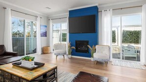 A spacious living room with a large TV mounted on a blue accent wall, a fireplace, and a view of the outdoors through large windows.