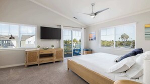 A bedroom with a large bed, a TV mounted on the wall, a ceiling fan, and a view of the outside through the windows.