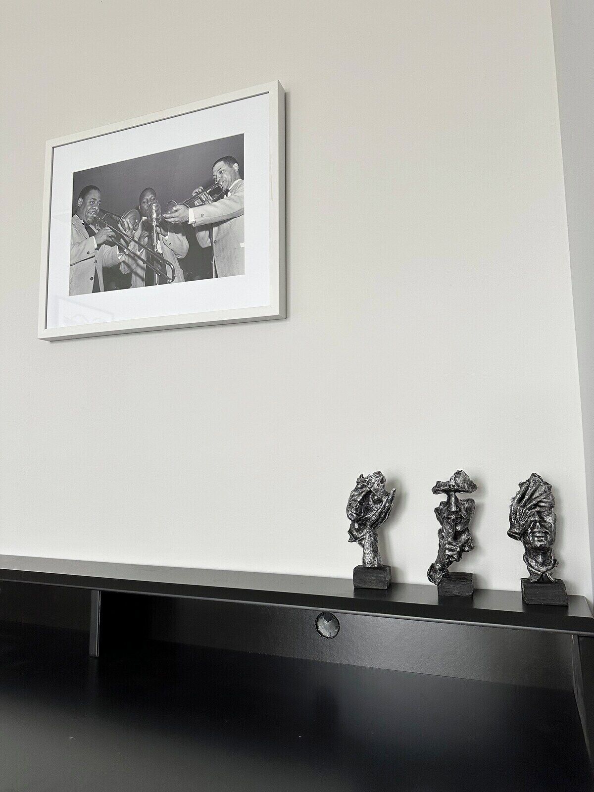 Fotografía en blanco y negro enmarcada de una banda de música de bronce sobre una pared, con tres esculturas metálicas en un estante negro debajo.