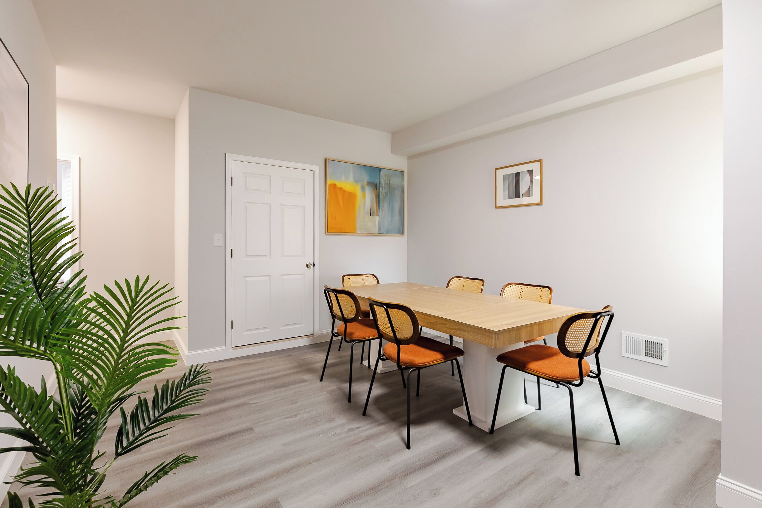 Uma sala de jantar com uma mesa de madeira e seis cadeiras, uma porta branca, um quadro e um vaso com planta.