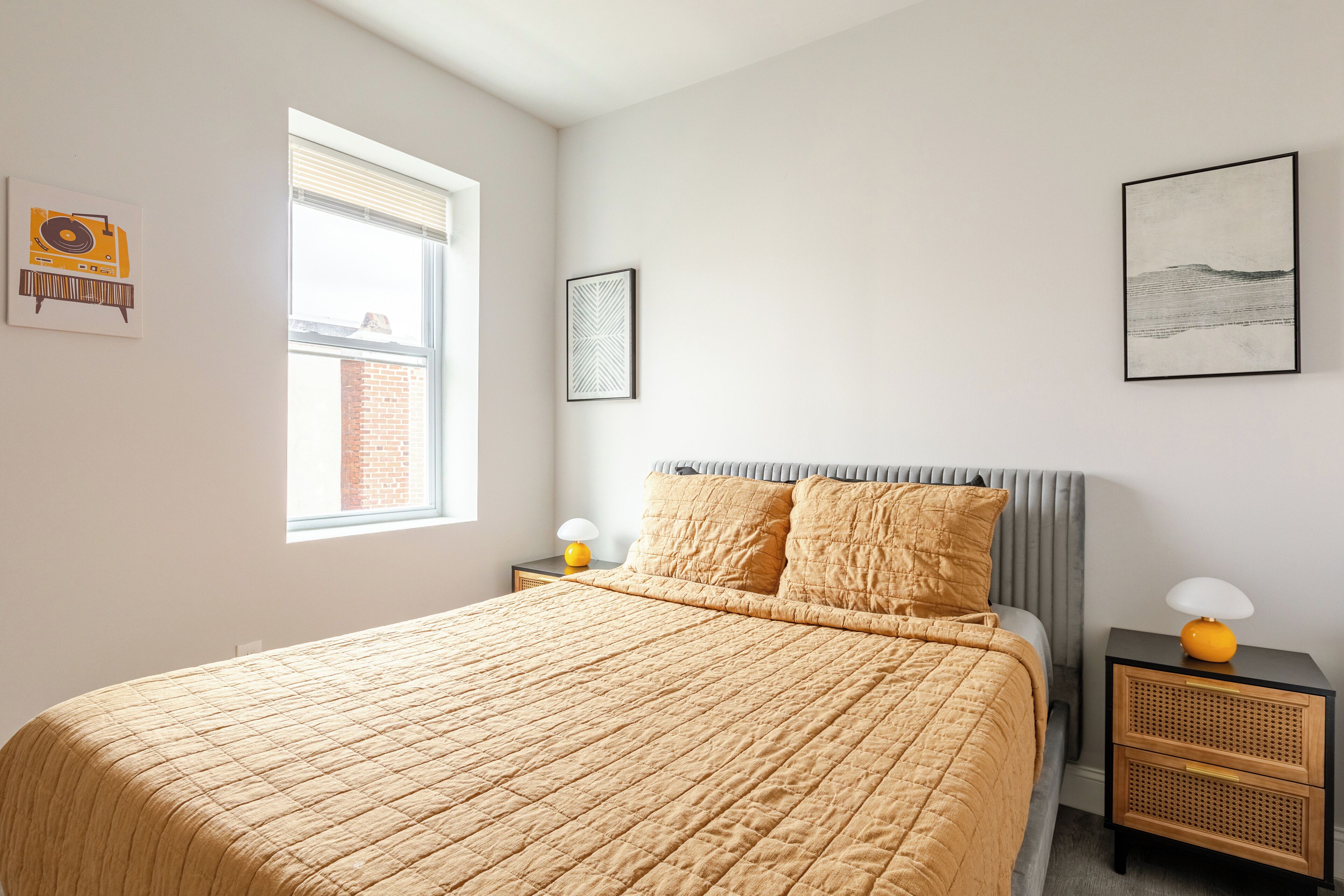 Une chambre à coucher avec un lit, deux tables de chevet, une fenêtre et des œuvres d’art accrochées aux murs.