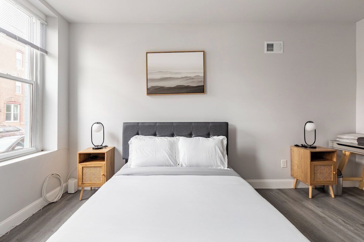 A modern bedroom with a large bed, two bedside tables with lamps, a wall-mounted picture, and a window with blinds.
