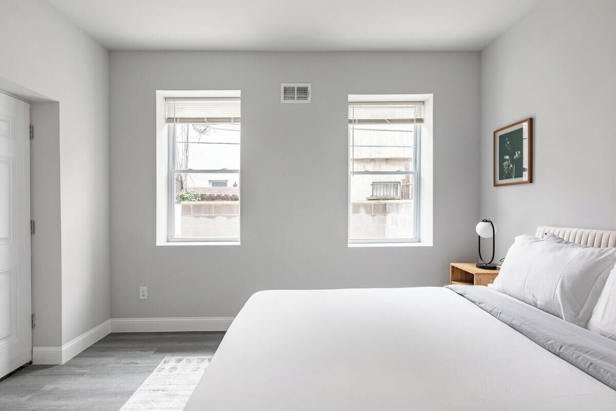 A bedroom with a large bed, two windows, a nightstand with a lamp, and a framed picture on the wall.