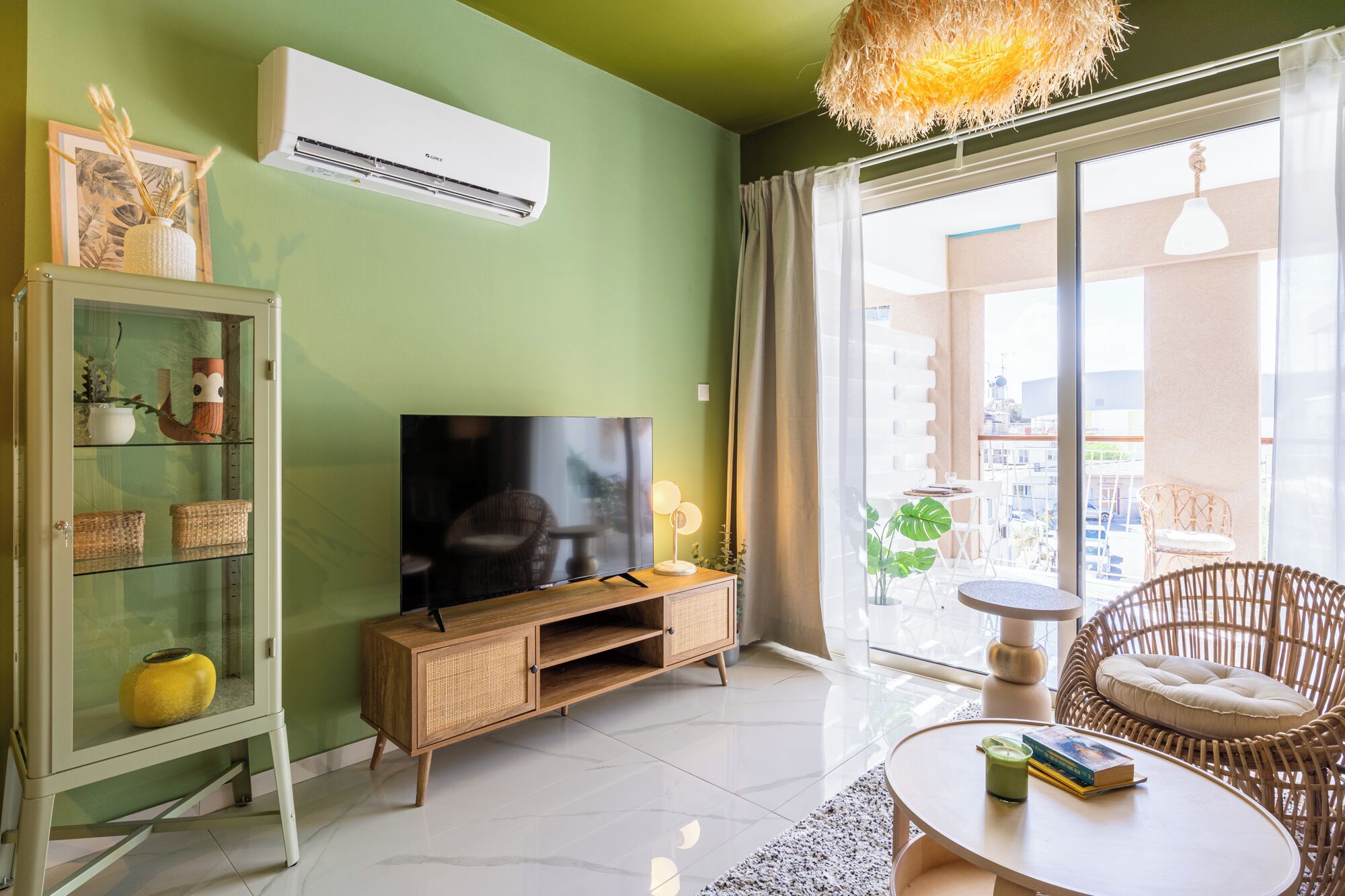 A living room with a TV, wicker furniture, a glass cabinet, and a balcony with plants.