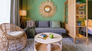Ein gemütliches Wohnzimmer mit grüner Wand, runder Wanddekoration aus Bändern, grauem Sofa, Korbstuhl, Holzregal und kleinem runden Tisch mit Büchern und Kerze.