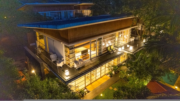 Una casa moderna de dos pisos con techo plano, ventanas grandes y un balcón con mobiliario exterior.