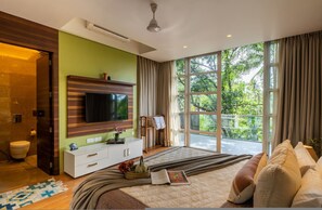 A modern bedroom with a large bed, a TV mounted on the wall, a window with curtains, and a view of greenery outside. - 4bhk w Pool 10 min to Baga Beach Villa Dahlia (Arpora)