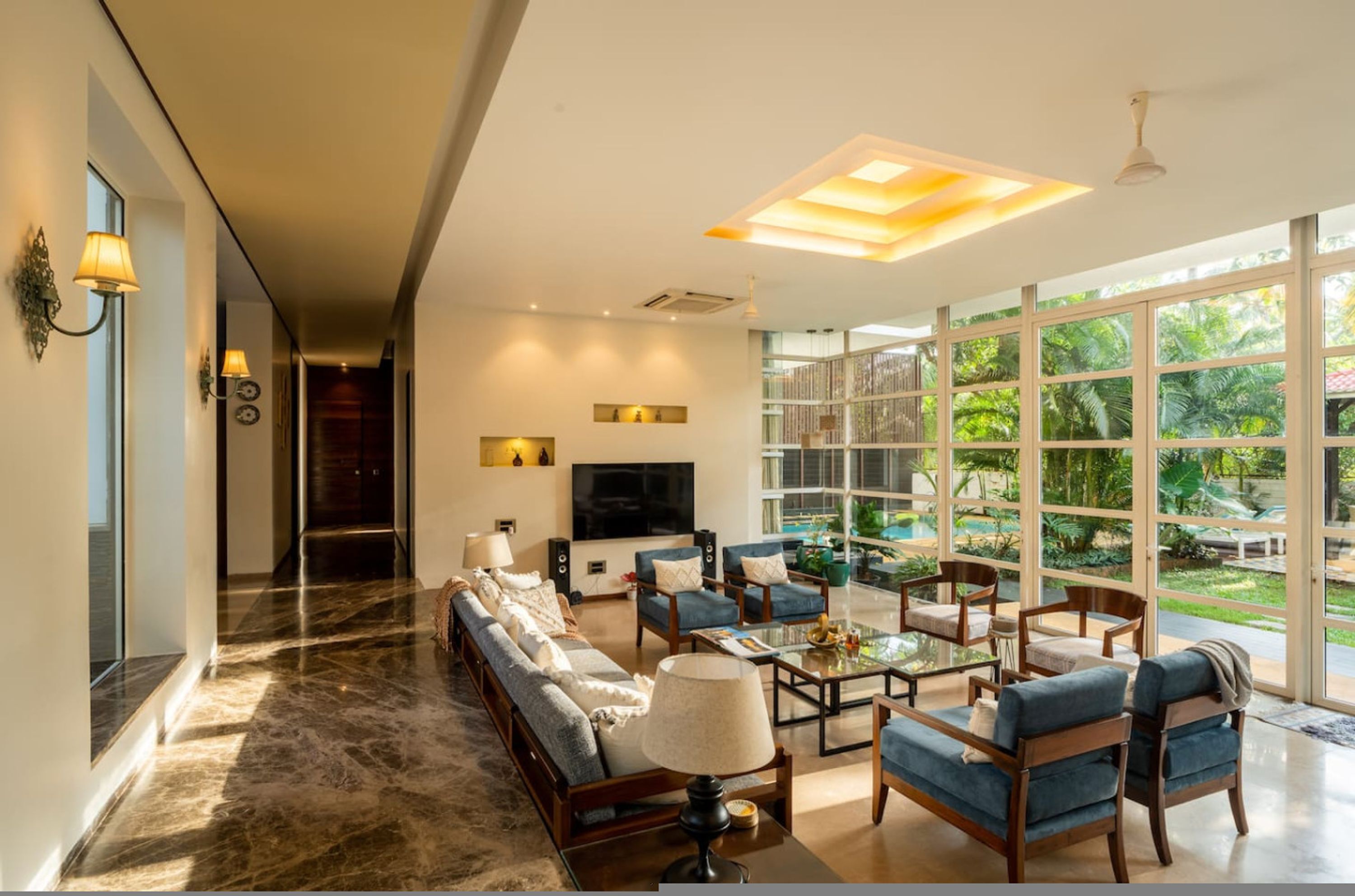 A spacious living room with a large sofa, armchairs, a coffee table, and a TV. There is a large window with a view of greenery outside.