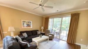 A living room with a dark grey sofa, a white coffee table, and a beige armchair. There is a ceiling fan, a window with curtains, and a painting on the wall. - Havens 1311 Beautiful Condo! (North Myrtle Beach)